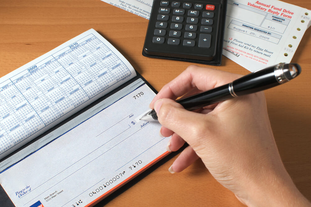 Woman’s Hand Writing a Check To Pay the Bills, With Calculator and an Invoice on the Desktop.